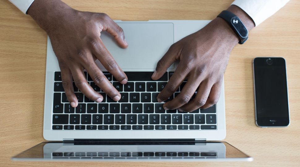 Black man typing on a laptop with a cell phone next to him.